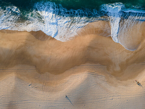 Aerial view of turquoise waves crashing onto the golden sands of Nazare's beach, creating a mesmerizing display of color and texture, Nazare, Leiria District, Portugal.