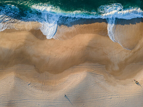 Aerial view of the meeting point where the dark turquoise ocean waves crash onto the golden sandy beach, creating a mesmerizing contrast, Nazare, Leiria District, Portugal.