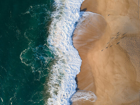 Aerial view of emerald waters clash with golden sands, where footprints trace paths along the shore, Nazare, Leiria District, Portugal.