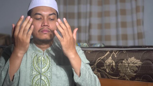 A devout Muslim man wearing a white skullcap and green clothes performing  prayer at home. He is raising his hands in supplication and wiping his face in a spiritual gesture. Peaceful religious moment