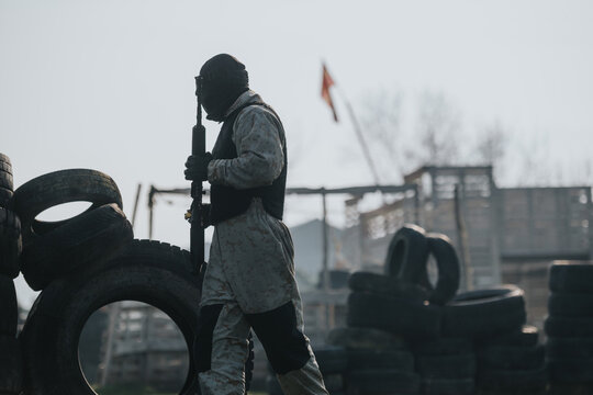 A masked paintball player in camouflage gear carries a marker amid stacked tires. The scene shows a rugged outdoor paintball field with obstacles and protective gear.