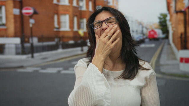 Hispanic woman wearing white blouse and glasses covering mouth with hand on city street; fatigue exhaustion.