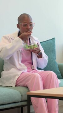 Vertical video: Starting break, clinician wearing pink scrubs eating salad with fork on teal sofa