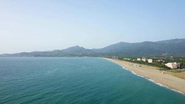 Wide aerial shot of Argeles beach with turquoise sea, sandy shoreline, and mountains in the background under a clear sky.