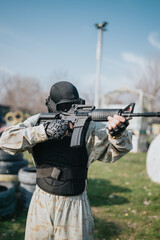 Fototapeta premium Paintball player in camouflage gear aims a rifle during an outdoor game. The scene shows a tactical athlete on a field with barriers and tires in the background.
