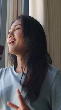 Vertical video: Raising red phone, blue-shirted woman singing by window with beige drape, gesturing