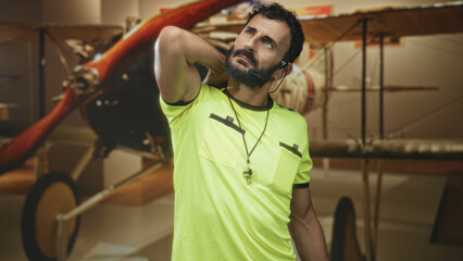 Man referee middle aged bearded hispanic with whistle and headset, hand behind neck stretch in a building with vintage biplane exhibit  tired reflection. © Krakenimages.com