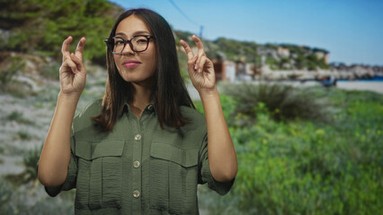 Obraz premium Young hispanic woman with glasses and brown hair making air quotes gesture in building near a green park and coastal dunes; playful skepticism.