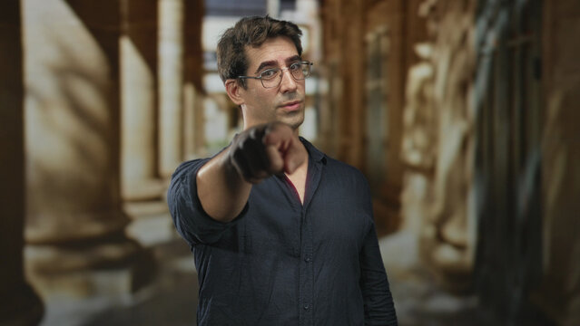 Man points finger forward while wearing glasses and a dark shirt in a columned building corridor; confidence challenge.