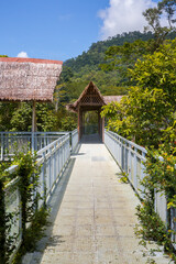 Fototapeta premium An elevated metal walkway in a jungle. The walkway is constructed with light-colored flooring and grey metal railings, featuring a small gabled hut with a thatched roof in the center-middle ground.