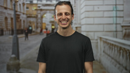Man smiling with visible teeth and long hair tied back on city street beside stone balustrade and lamppost while wearing black t shirt; joy confidence.