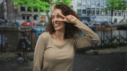 Fototapeta premium Woman smiling makes peace sign with fingers by eye on amsterdam street by canal and parked bicycles while laughing and looking sideways; playful joy.