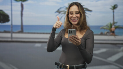 Woman wearing glasses and jeans tapping a smartphone with both hands while smiling on a seaside street lined with palm trees and a road; joyful surprise.