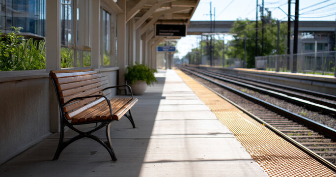 Empty wooden bench on a sunny train station platform with railway tracks and modern urban surroundings in the background
