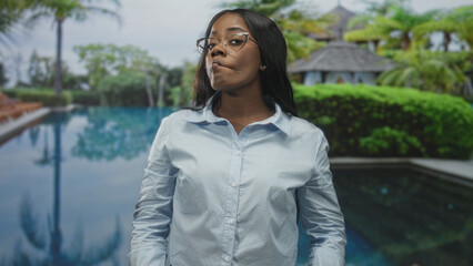 Woman wearing glasses pursing lips with a slight head tilt by a resort pool and gazebo, poolside...