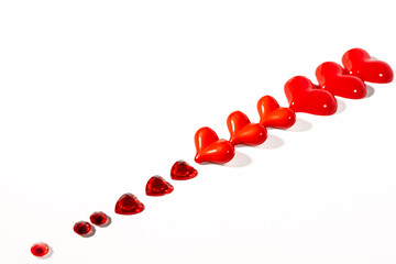 Glossy red and orange heart-shaped ornaments aligned diagonally on a clean white background, with small faceted red heart gemstones forming a leading trail. Minimal romantic composition with copy.