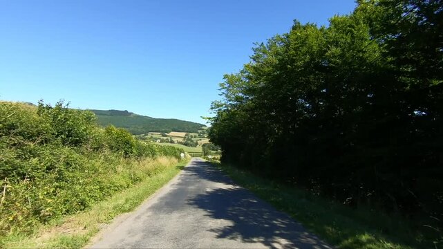 Empty country road winding through the green hills and fields of the Morvan region under a clear blue sky. Peaceful rural landscape with lush vegetation and distant hills.
