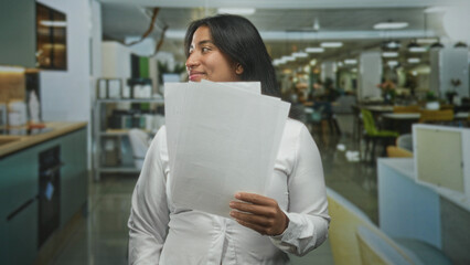 Fototapeta premium Young hispanic woman holding a stack of papers, speaking with her hand visible, in a modern office building with desks and seating; public speaking confidence.