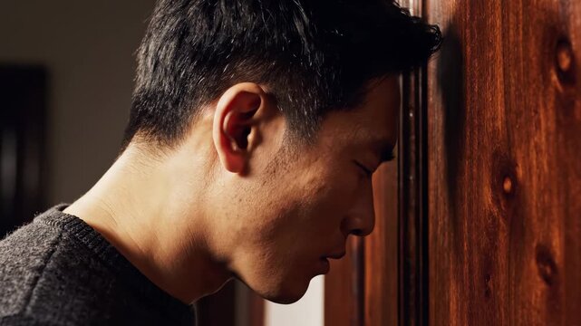 In a muted space filled with rich wood tones, a man leans against the wall with his forehead gently resting, lost in deep thought. The ambiance invites contemplation and personal reflection.