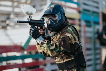 A paintball player in camouflage gear wears a full-face mask and helmet while aiming a marker. The scene shows an outdoor arena with obstacles as the player takes a precise shot. © qunica.com