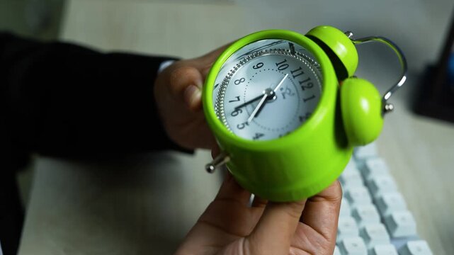 Close-up of hands holding a bright green twin-bell alarm clock over a keyboard, office desk background shallow depth of field