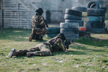 Fototapeta premium Two camo-clad players crouch and lie prone with paintball markers during a field game. The scene shows stacked tires used as cover on a grassy course.