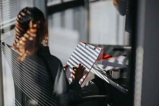 Person with clipboard studies notes in a sunlit office setting. Window blinds cast striped shadows over desk and coffee mug.