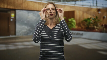 Fototapeta premium Woman in striped shirt holding eyeglasses and adjusting frames while standing in a modern building lobby; thoughtful focus.