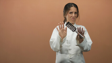 Woman chef in white coat holding metal spatula with hand on chest and a wincing expression in studio  hesitation concern reflection solitude. © Krakenimages.com