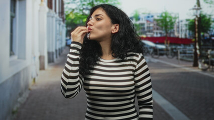 Fototapeta premium Young arab woman holding fingers to lips in a kiss gesture on a city street, striped shirt and curly hair visible; playful.
