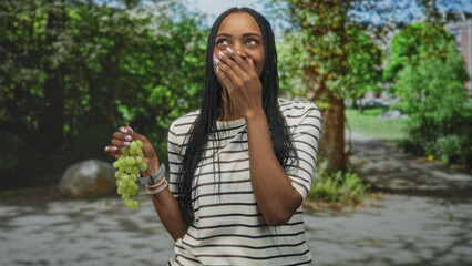 Fototapeta premium Woman holding green grapes, hand covering mouth in a laughing gesture, wearing a striped shirt on a street lined with trees and stone path; joyful surprise.