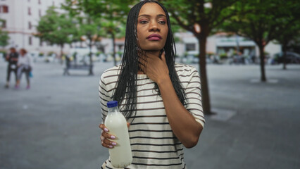 Fototapeta premium Woman holding a milk bottle by the cap with one hand, touching cheek and neck with the other, striped shirt and braids visible on a street plaza; quiet contemplation.