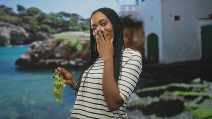Fototapeta premium Woman holding a bunch of green grapes while covering her mouth with hand in a studio with seaside backdrop, laughing and smiling candidly; joy.