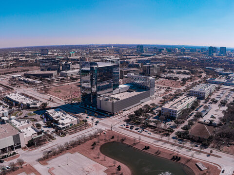 Aerial view of modern buildings casting long shadows under a clear blue sky, with a small pond reflecting the day's light, Frisco, Texas, United States.