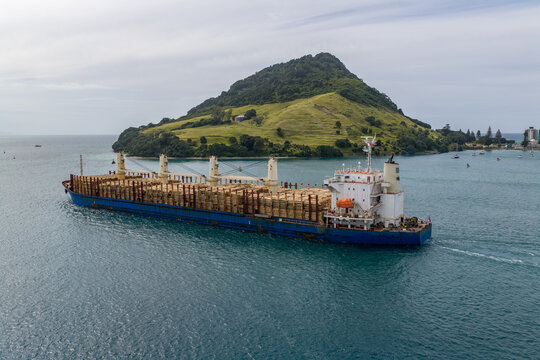 Aerial view of a cargo ship laden with logs sails past Mauao (Mount Maunganui) under a soft sky, Tauranga, Bay of Plenty Region, New Zealand.