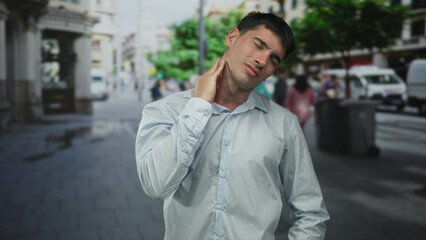 Fototapeta premium Hispanic man in white shirt grimacing with hand on neck on sunlit street near city building; discomfort.