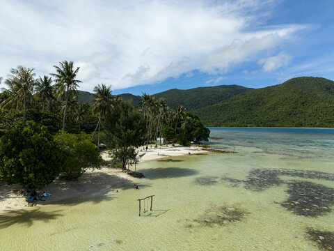 Aerial view of Bobby Beach's turquoise waters meeting white sands under a canopy of palm trees, framed by verdant hills, Karimunjawa, Central Java, Indonesia.
