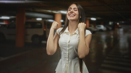 Fototapeta premium Teenage girl in a white dress with braces, fists clenched and smiling in an underground parking garage building; celebration joy relief.