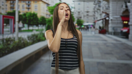 Fototapeta premium Teen girl touching her face with right hand while looking uncertain, long straight hair and striped top, standing alone on an urban street; contemplation.