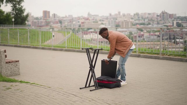 black street musician setting up keyboard outdoors city park, unpacking case, mounting stand, plugging cables, adjusting headphones, testing synth patches, looping beatmaker sequence, focused