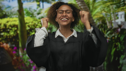 Fototapeta premium Woman in black robe with fists raised smiling broadly and wearing glasses in forest with lush green plants; academic achievement celebration.
