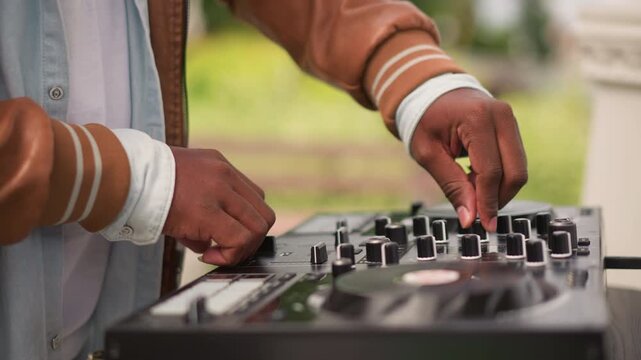 park black DJ hands adjusting knobs on controller during sunlit outdoor set casual jacket and wristband, portable mixer and laptop, precise mixing and loop triggering, upbeat festival energy, live