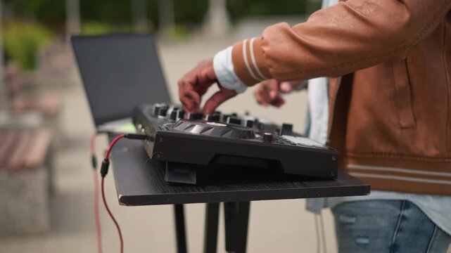 dj hands on mixer outdoor park closeup showing fingers adjusting knobs, laptop beside controller, red cable hanging, casual jacket and jeans, benchlined walkway, daytime focused tactile vibe, street