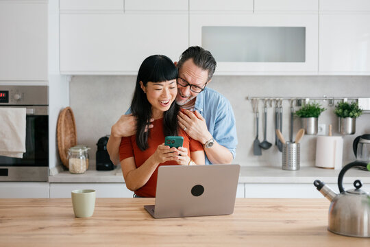 Happy couple buying online together using smartphone in kitchen
