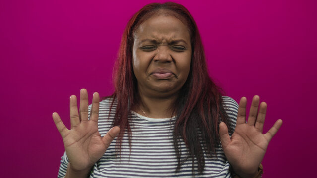Woman in striped shirt face grimaces and eyes closed with raised hands in frontlit pink studio; disgust.