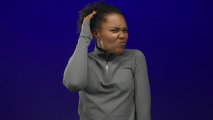 Young african american woman scratching head gesture in studio on vivid blue wall wearing grey...