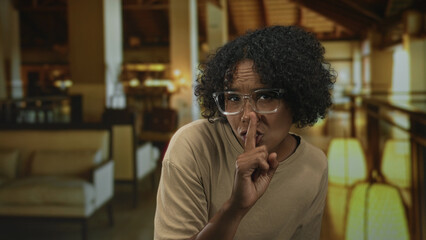 Young woman wearing glasses with curly hair stands with finger to lips for silence in building  mystery. © Krakenimages.com