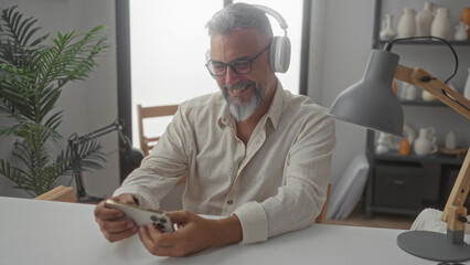 Man holding smartphone and smiling while wearing headphones and glasses at studio desk, hands on...