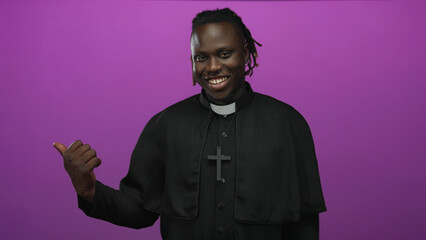 Obraz premium Young african man priest pointing thumb to side wearing cross necklace in purple studio; welcoming joy community.