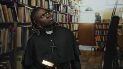 Man priest holding an illuminated book in one hand, wearing a clerical collar and large cross, looking upward among tall bookshelves inside a library building  faith contemplation. © Krakenimages.com